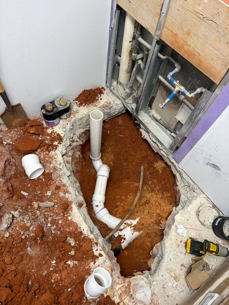 A commercial restroom slab cut open with new white PVC drain line run through the red clay beneath it and the wall opened to expose the existing supply piping