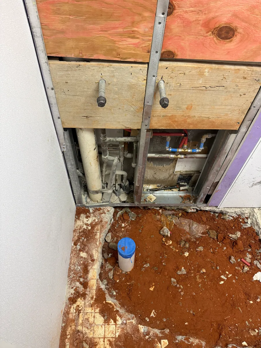 A plumbing chase opened in a commercial restroom wall with copper and PEX supply lines and a PVC vent, and a capped PVC stub-up standing in the excavated slab below