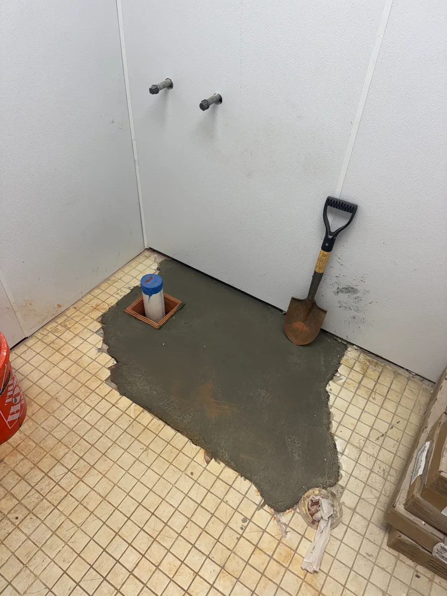 A restroom slab patched with fresh concrete around a capped PVC stub-up, with two capped supply stubs coming out of the finished wall above it